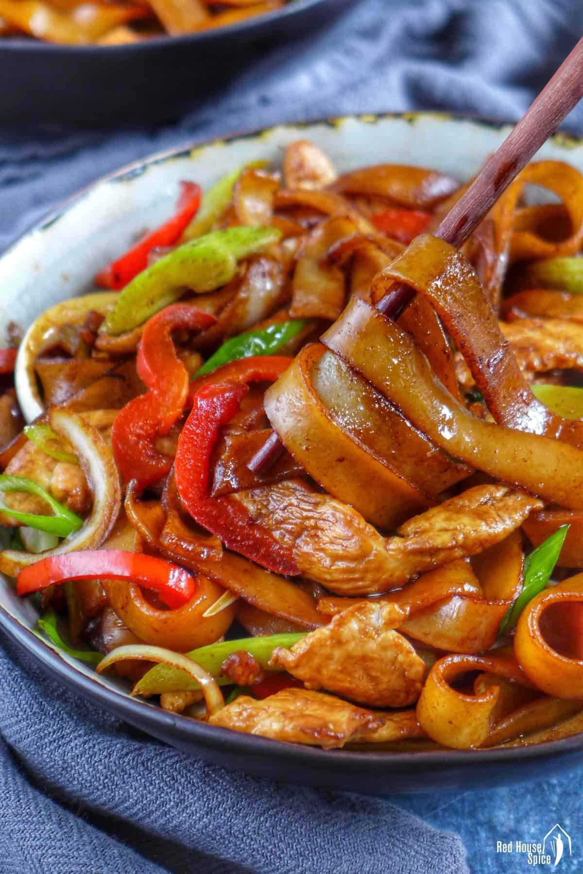 chopsticks picking up wide rice noodles cooked with chicken and vegetables.