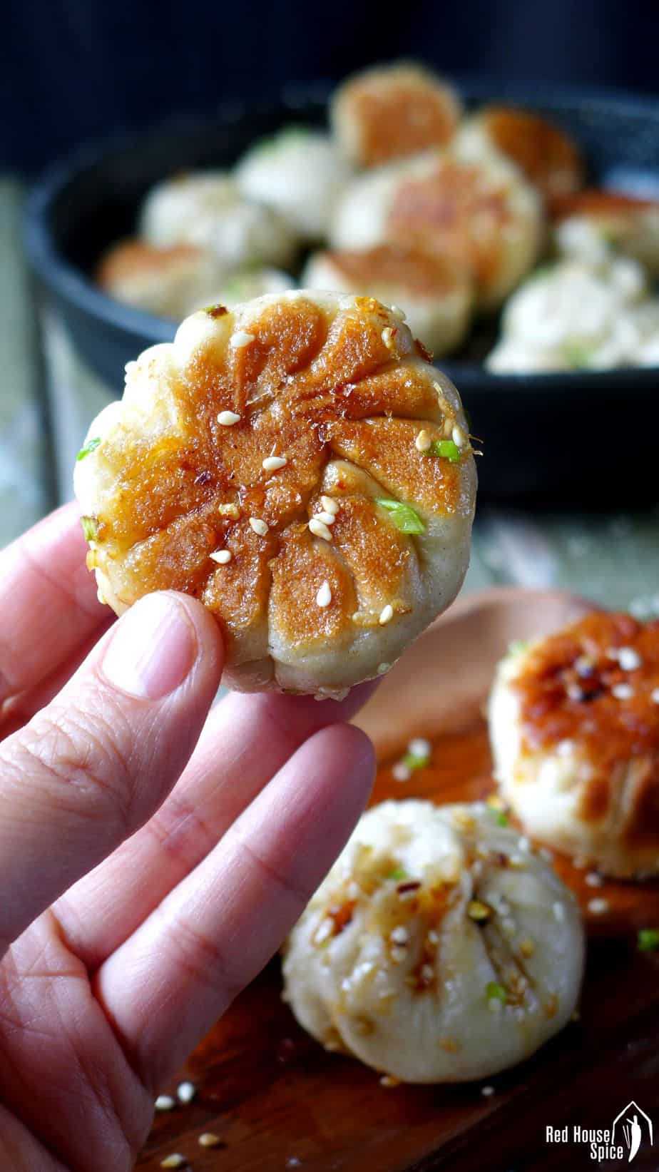 A hand holding a Sheng Jian Bao (pan-fried pork buns) with a golden, crispy crust.